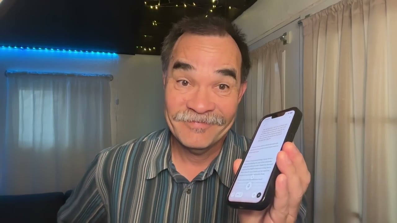 Mash holding up a phone with Claude open, mid-conversation, in a home office with blue lights and curtains.